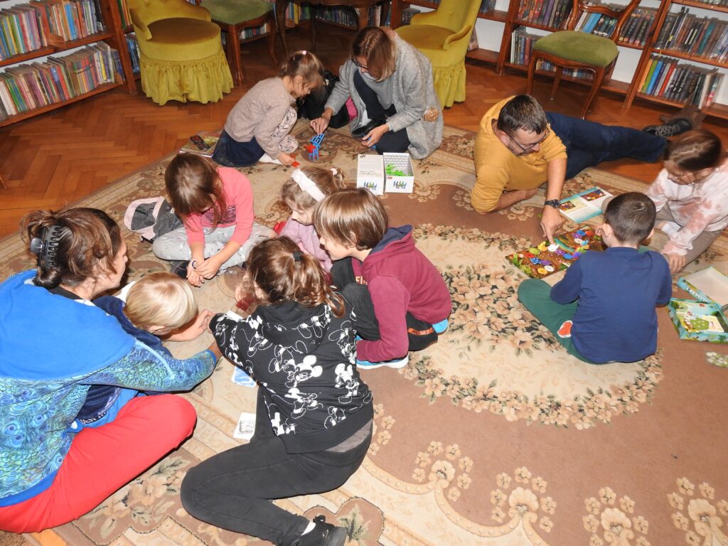 Jedenaście osób siedzi na dywanie. Tworzą trzy grupy. Przed każdą z nich rozłożona jest gra planszowa.