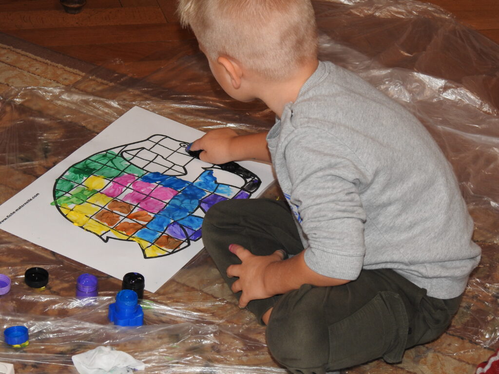 Jedno dziecko siedzi na dywanie pokrytym folią. Przed nim znajduje się kartka A3, na której znajduje się nadrukowany szablon słonia podzielonego na kwadraty. Dziecko maluje trąbę słonia palcem umoczonym w czarnej farbie. Większość postaci słonia jest już pokolorowana. Obok pracy znajdują się otwarte pojemniczki z farbami.