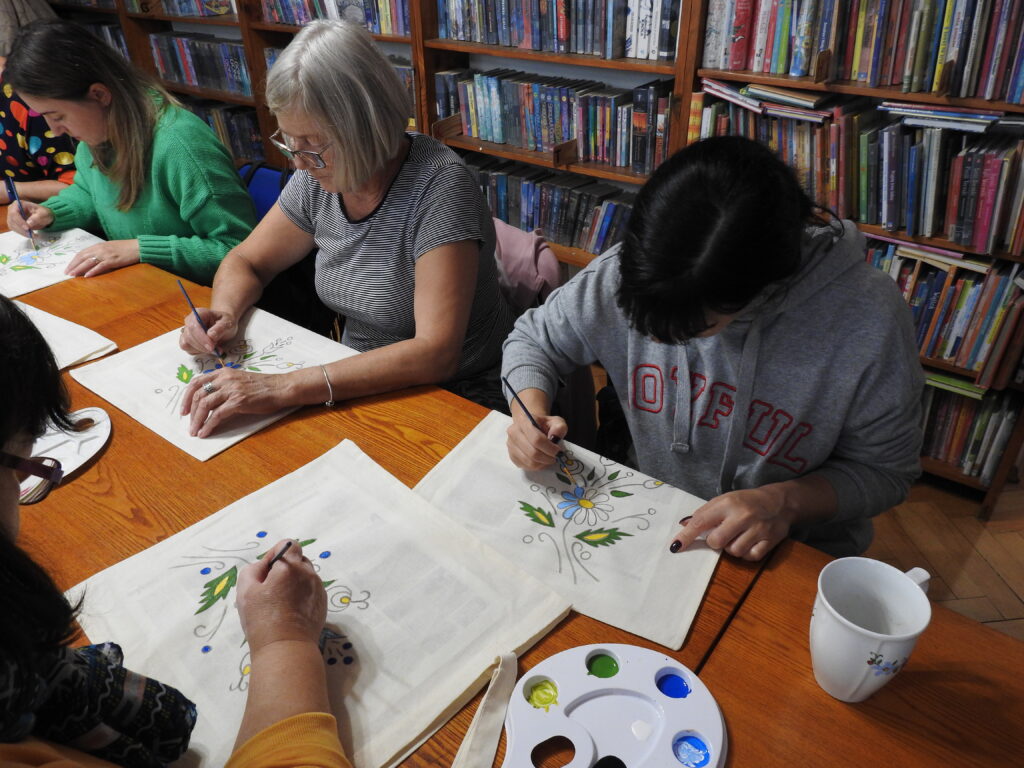 4 kobiety siedzą przy stole, na którym znajdują się płócienne torby oraz paleta z farbami. Kobiety malują pędzelkami na torbach. 