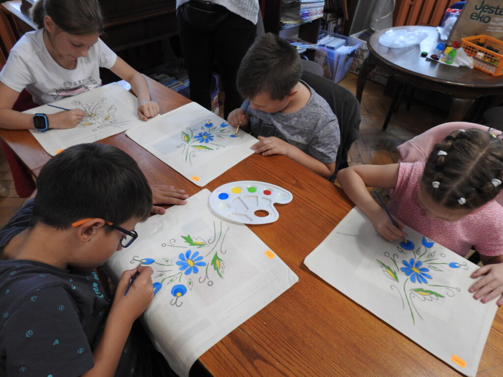 Czworo dzieci siedzi przy stole, na którym znajdują się płócienne torby oraz paleta z farbami. Dzieci kolorują kwiatowy kaszubski wzór na torbach.