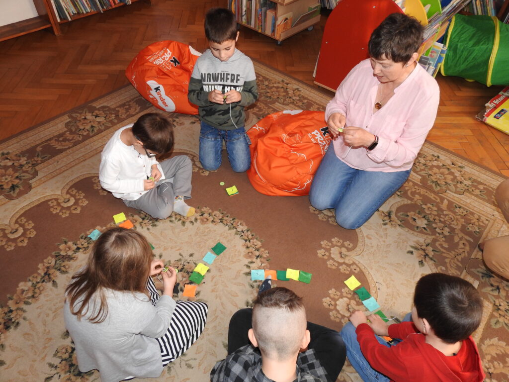 Pięcioro dzieci oraz jedna osoba dorosła siedzą lub klęczą na dywanie. Przed nimi leżą kolorowe kawałki materiału w kształcie kwadratów. Wszyscy nakładają guziki na sznurki.