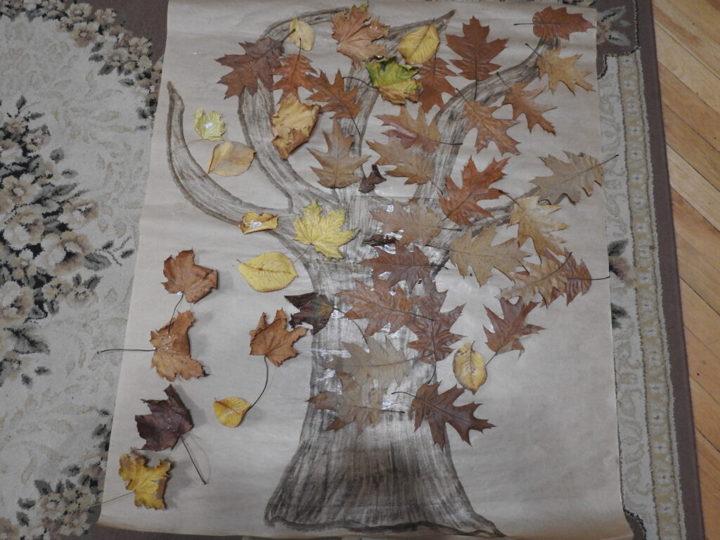 Na dywanie leży duży arkusz szarego papieru z namalowanym drzewem i przyklejonymi do niego prawdziwymi liśćmi różnego koloru i kształtu.