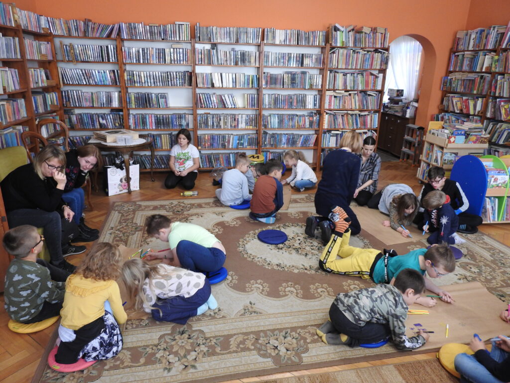 Grupa dzieci siedzi lub leży na dywanie Są podzieleni w grupy. Kredkami lub mazakami malują na dużych, szarych arkuszach papieru. Dwie osoby dorosłe siedzą z boku na fotelach, a dwie klęczą pomiędzy dziećmi obserwując jeden z rysunków.