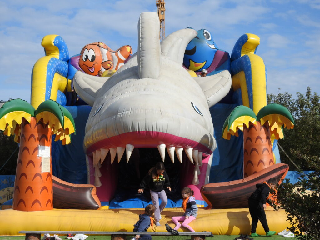 Troje dzieci stoi lub siedzi na przedniej części wielkiego dmuchańca w kształcie rekina. Jedno dziecko idzie obok dmuchańca. 