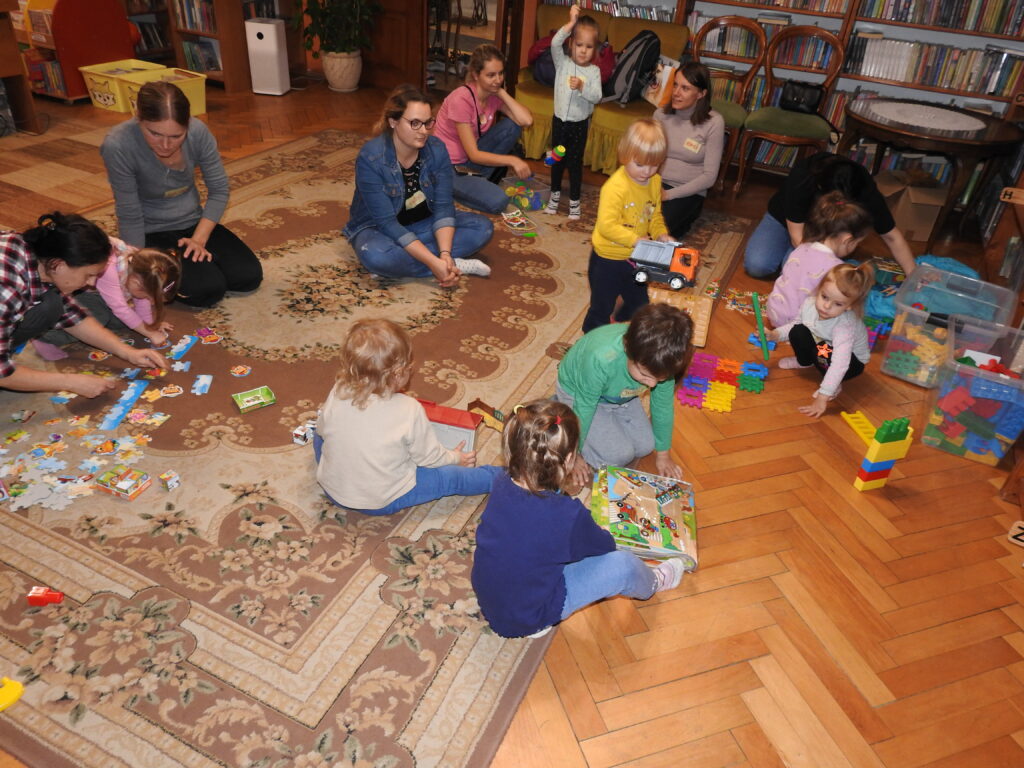 Grupa dzieci oraz osób dorosłych bawi się na dywanie różnymi zabawkami – puzzlami, klockami, samochodami.