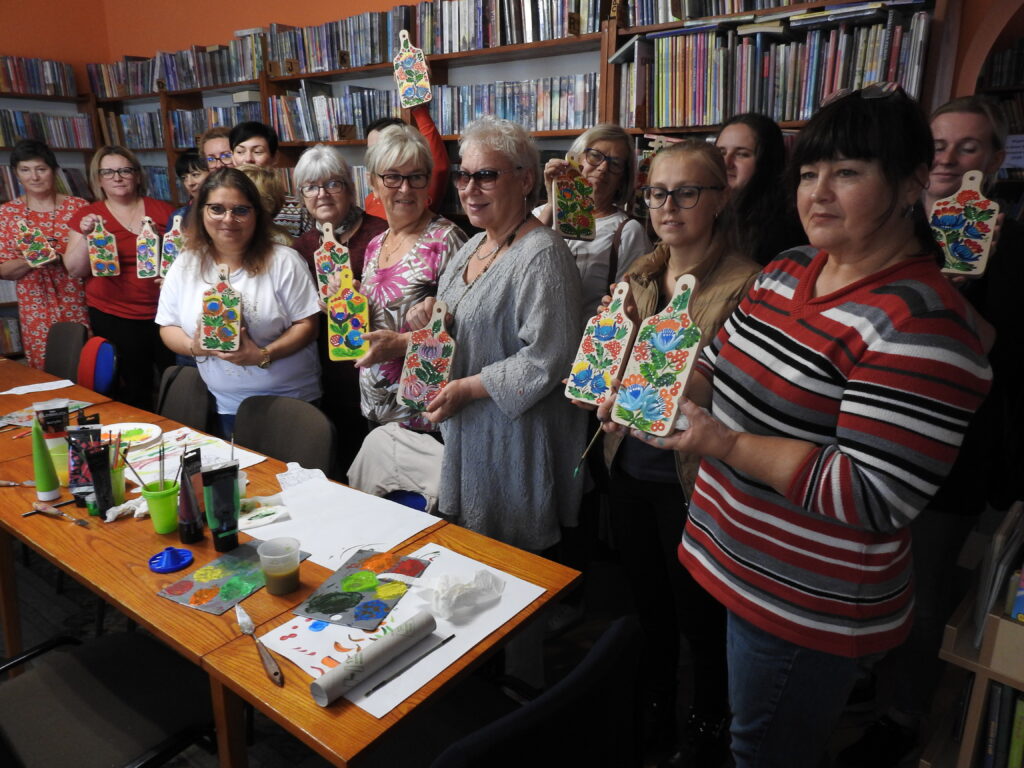 Duża grupa kobiet stoi trzymając w ręce drewniane deski z namalowanym kwiatowym wzorem. Pomiędzy nimi stoi Marta Zwarycz. Przed nimi stół z farbami, kartkami, pojemnikami z wodą. W tle regały z książkami.