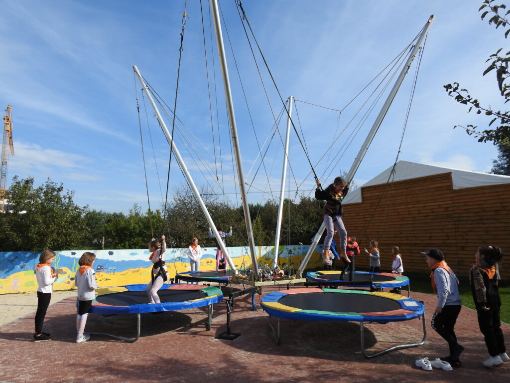 Czworo dzieci znajduje się na trampolinach. Są przyczepieni do specjalnej uprzęży zapewniającej bezpieczeństwo podczas skoków. Kilkoro dzieci stoi w kolejce przed każdą z trampolin. W tle ogrodzenie z rysunkiem przedstawiającym plażę oraz fragment niewielkiego, drewnianego budynku.
