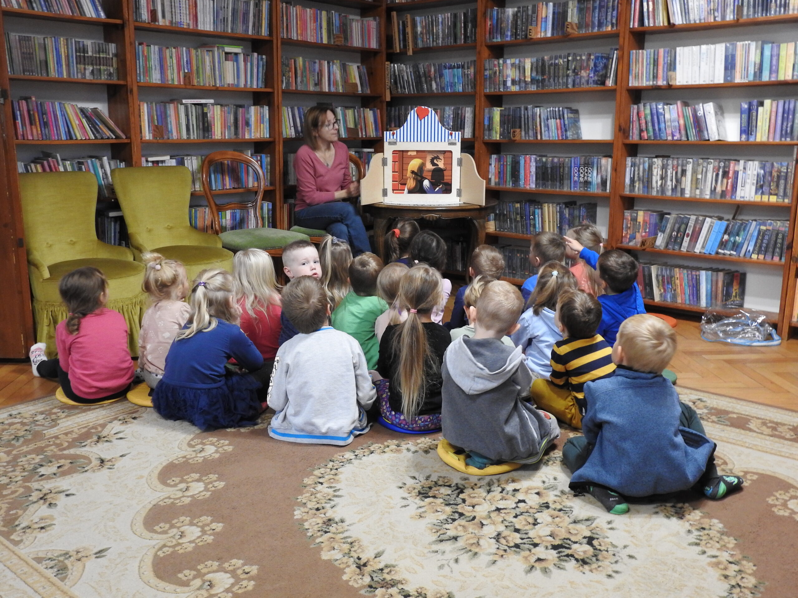 Bibliotekarka czyta siedzącym na dywanie dzieciom Legendę o smoku wawelskim. Historia przedstawiona jest za pomocą teatru kamishibai. Na karcie narracyjnej widzimy chłopca z zarzuconym na ramię tobołkiem, który czyta informację o smoku.