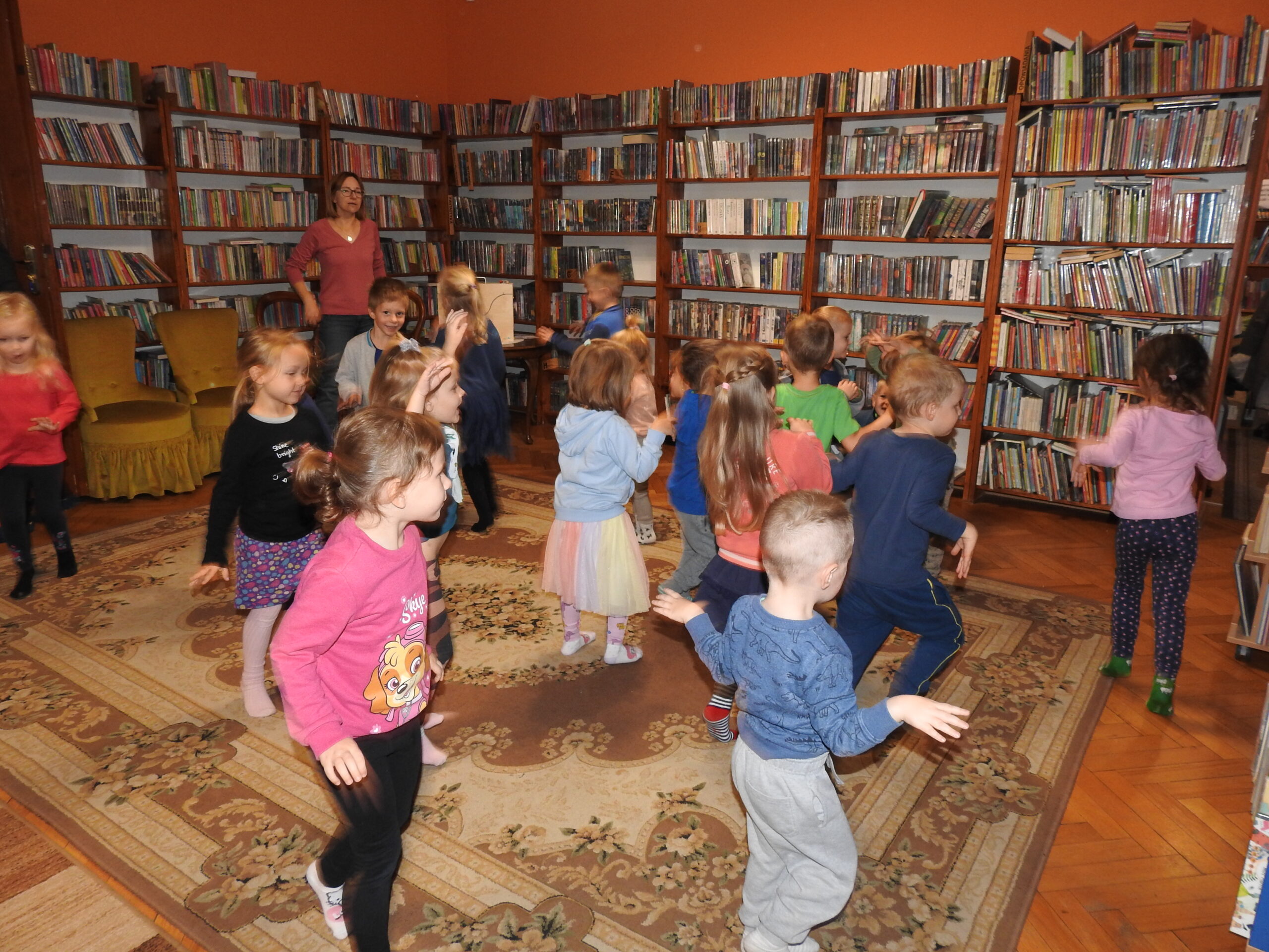 Grupa dzieci bawi się z bibliotekarką w smoka. Dzieci biegają z uniesionymi do góry rękoma. 