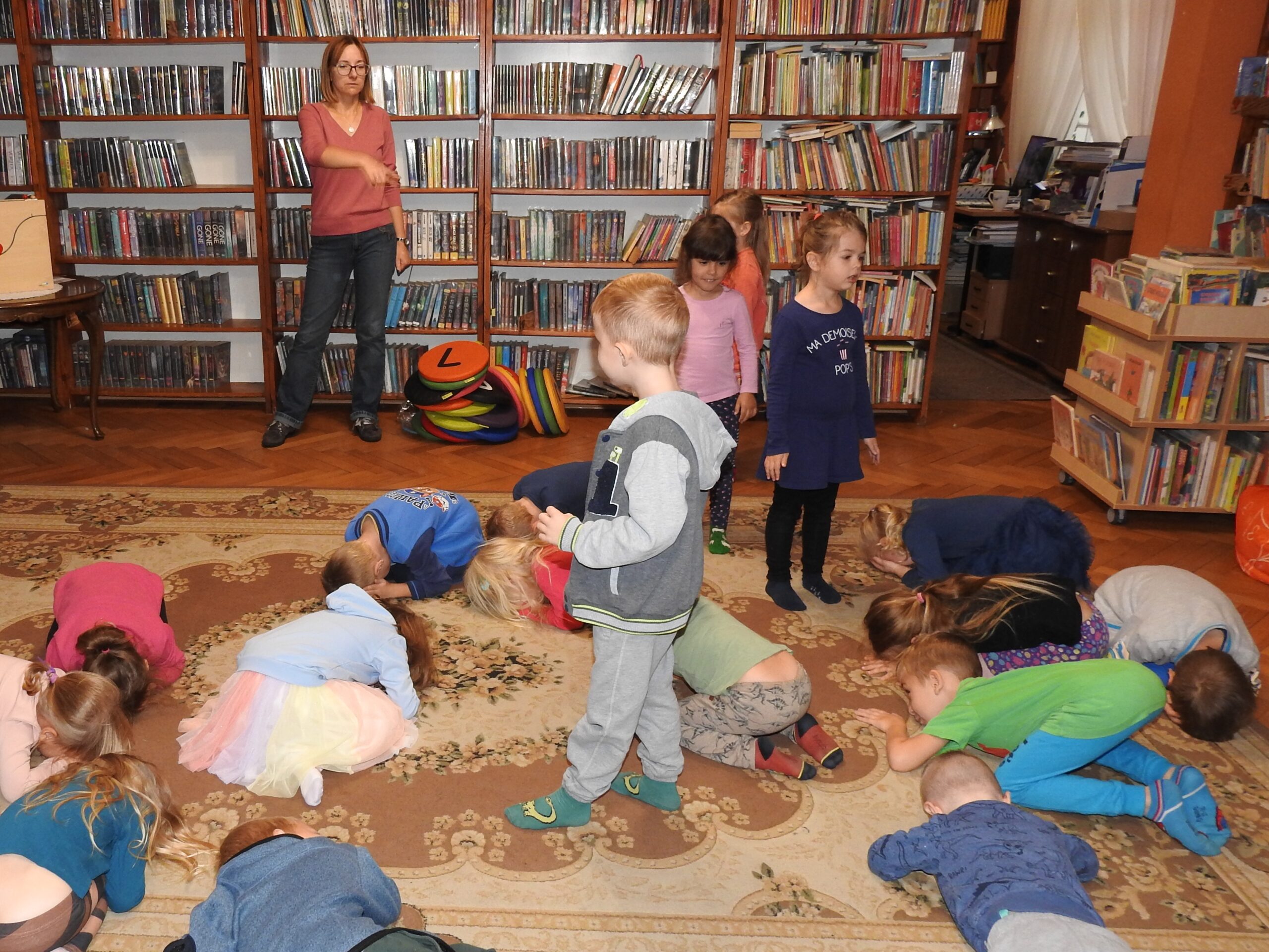 Dzieci bawią się z bibliotekarką w smoka. Chłopiec i 3 dziewczynki stoją. Pozostałe dzieci leżą w klęczkach na dywanie, z twarzami zwróconymi w dół.