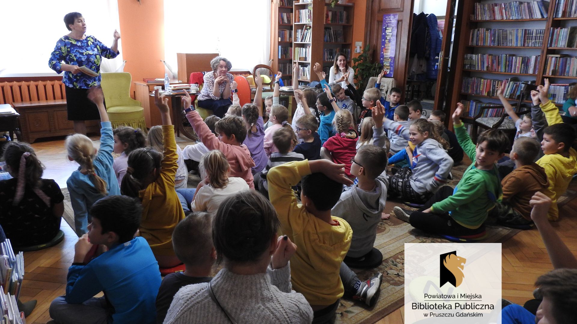 Pani Renata Piątkowska stoi. W jednej ręce trzyma książkę, a drugą rękę ma uniesioną lekko w górę. Obok, na fotelu, siedzi pani Malwina Kożurno. W stronę kobiet zwrócona jest duża grupa dzieci siedząca na dywanie. Wiele dzieci ma uniesioną do góry rękę.