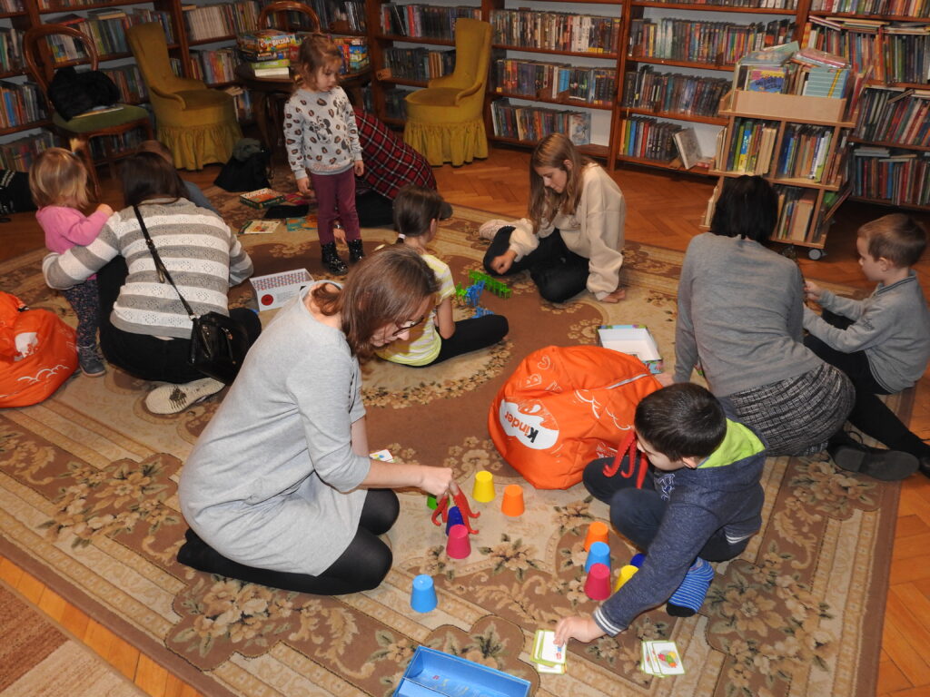 Cztery osoby dorosłe oraz siedmioro dzieci siedzą na dywanie podzieleni na kilka grup. Przed każdą grupą rozłożona jest gra planszowa.
