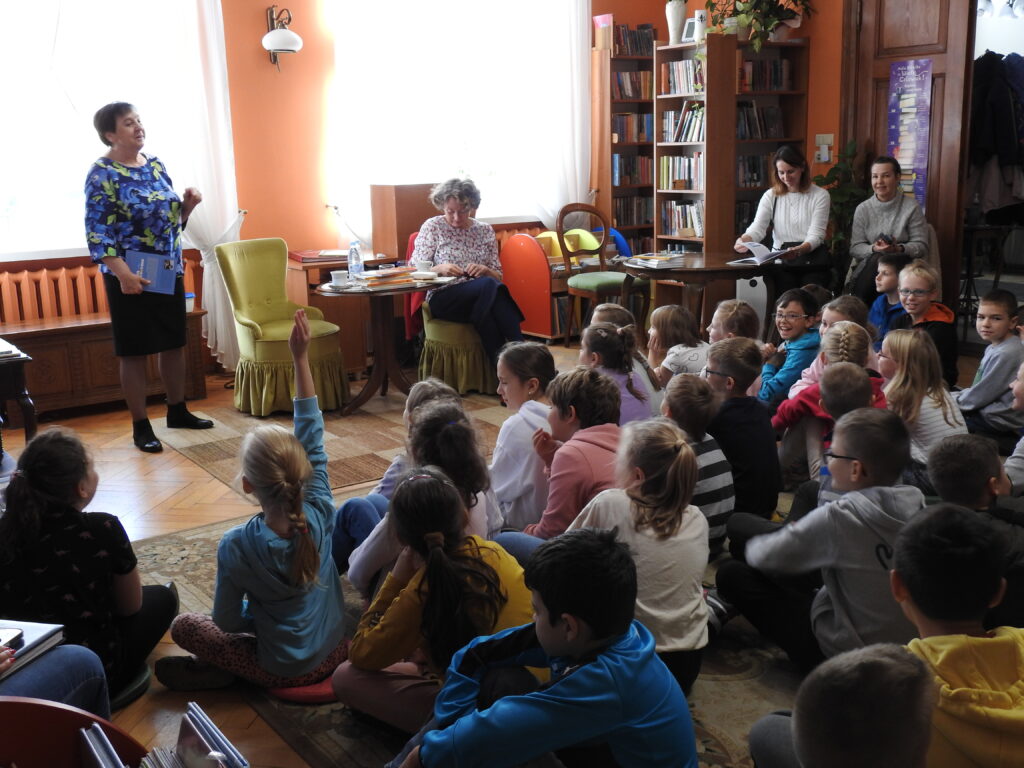 Pani Renata Piątkowska stoi. W jednej ręce trzyma książkę. Obok, na fotelu, siedzi pani Malwina Kożurno. W stronę kobiet zwrócona jest duża grupa dzieci siedząca na dywanie. Po prawej stronie, przy stole, na krzesłach siedzą dwie osoby dorosłe.
