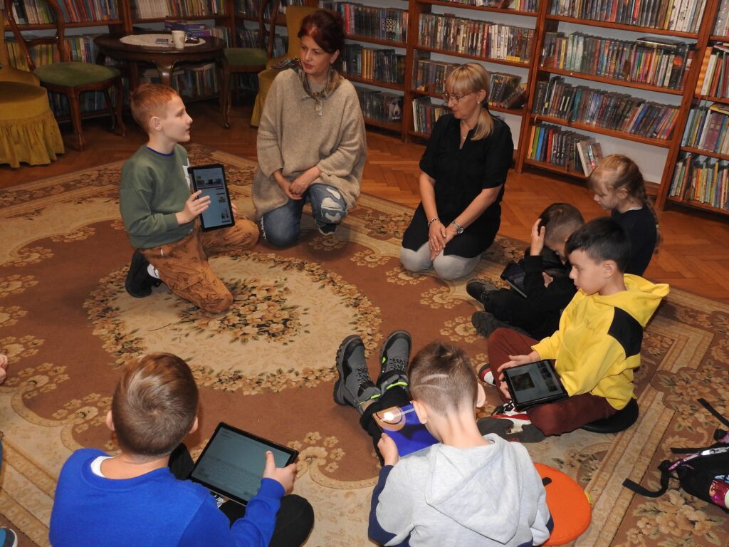 Sześcioro dzieci oraz dwie osoby dorosłe siedzą w półokręgu na dywanie. Dzieci trzymają w rękach tablety. Jedno z dzieci pokazuje coś na tablecie osobom dorosłym.