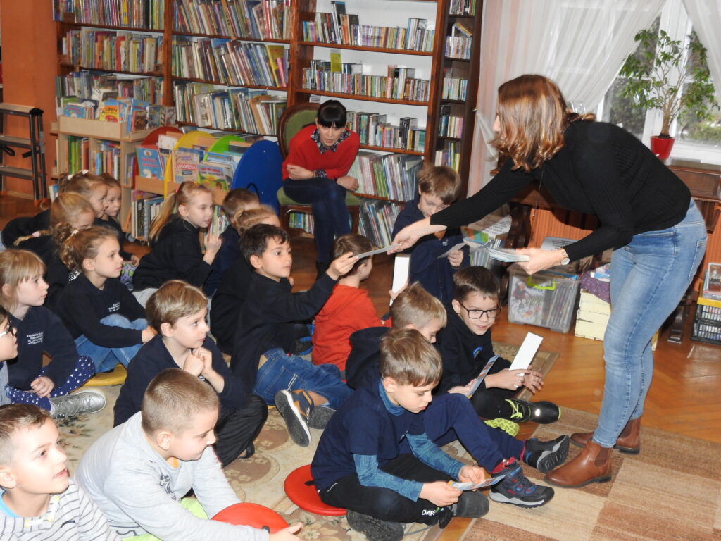 Magda Bielicka wręcza zakładki do książek siedzącym przed nią dzieciom.  Z tyłu, na krześle siedzi osoba dorosła. W tle regały z książkami.