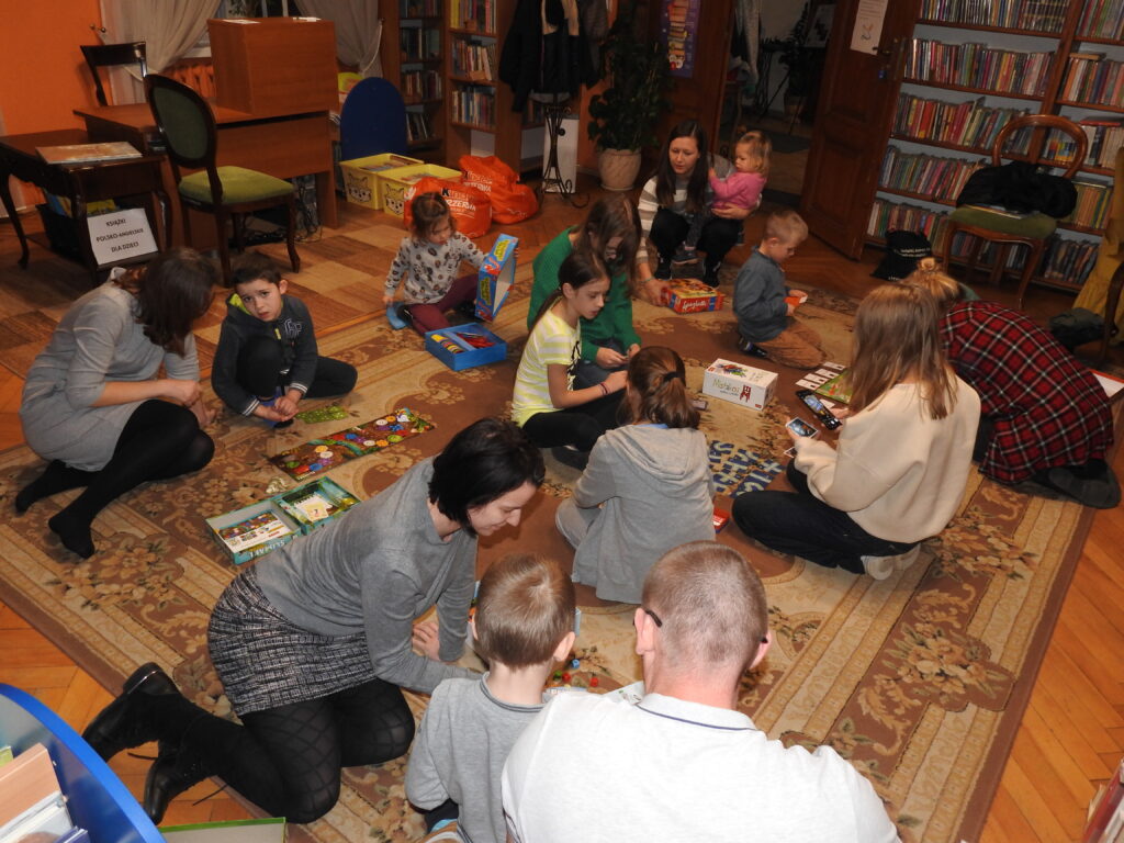 5 osób dorosłych oraz 9 dzieci siedzi na dywanie. Są podzieleni na kilka grup. Przed każdą z nich rozłożona jest gra planszowa. 