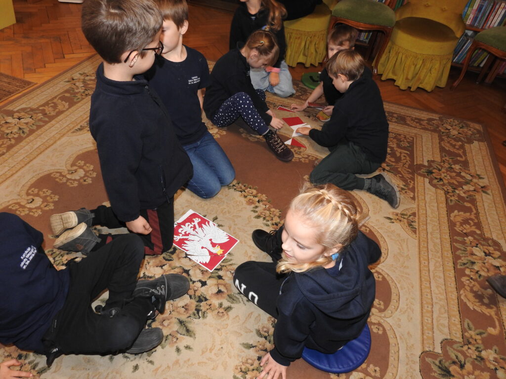Kilkoro dzieci siedzi lub klęczy na dywanie. Przed nimi znajdują się wydrukowane dwa godła Polski pocięte na kawałki i tworzące puzzle. Dzieci je układają.