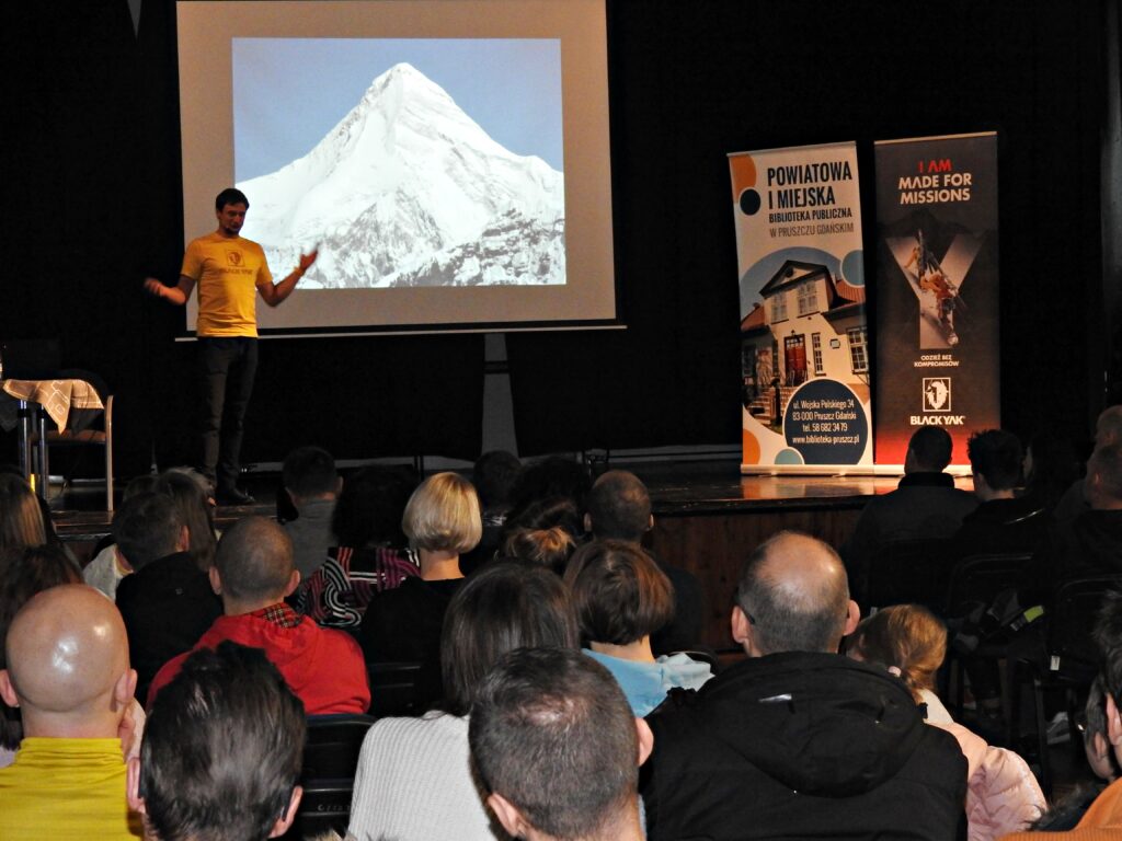 Adam Bielecki stoi na scenie z rozłożonymi rękami. Za nim ekran ze zdjęciem zaśnieżonego szczytu górskiego. Naprzeciw sceny siedzi duża grupa ludzi.