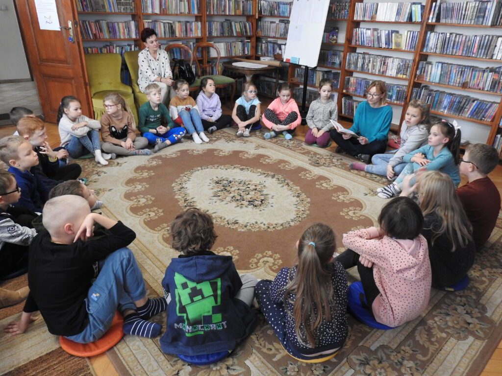 Duża grupa dzieci oraz jedna osoba dorosła siedzą w kole na dywanie. Obok, na fotelu, siedzi kobieta. W tle regały z książkami.