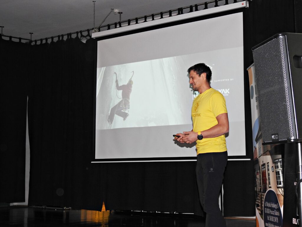 Adam Bielecki stoi obok ekranu, na którym wyświetlony jest człowiek podczas wspinaczki górskiej.