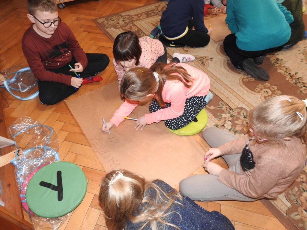 Pięcioro dzieci siedzi na dywanie wokół arkusza szarego papieru. Jedno z dzieci pisze na arkuszu mazakiem.