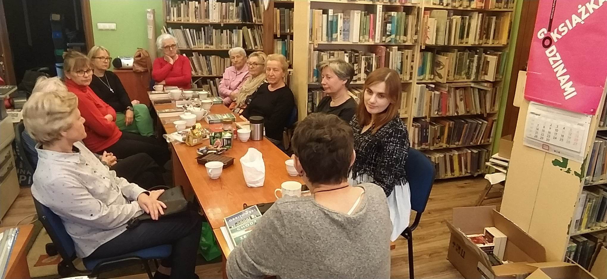Jedenaście kobiet siedzi przy stole, na którym stoją filiżanki z kawą, ciasteczka oraz leżą książki.