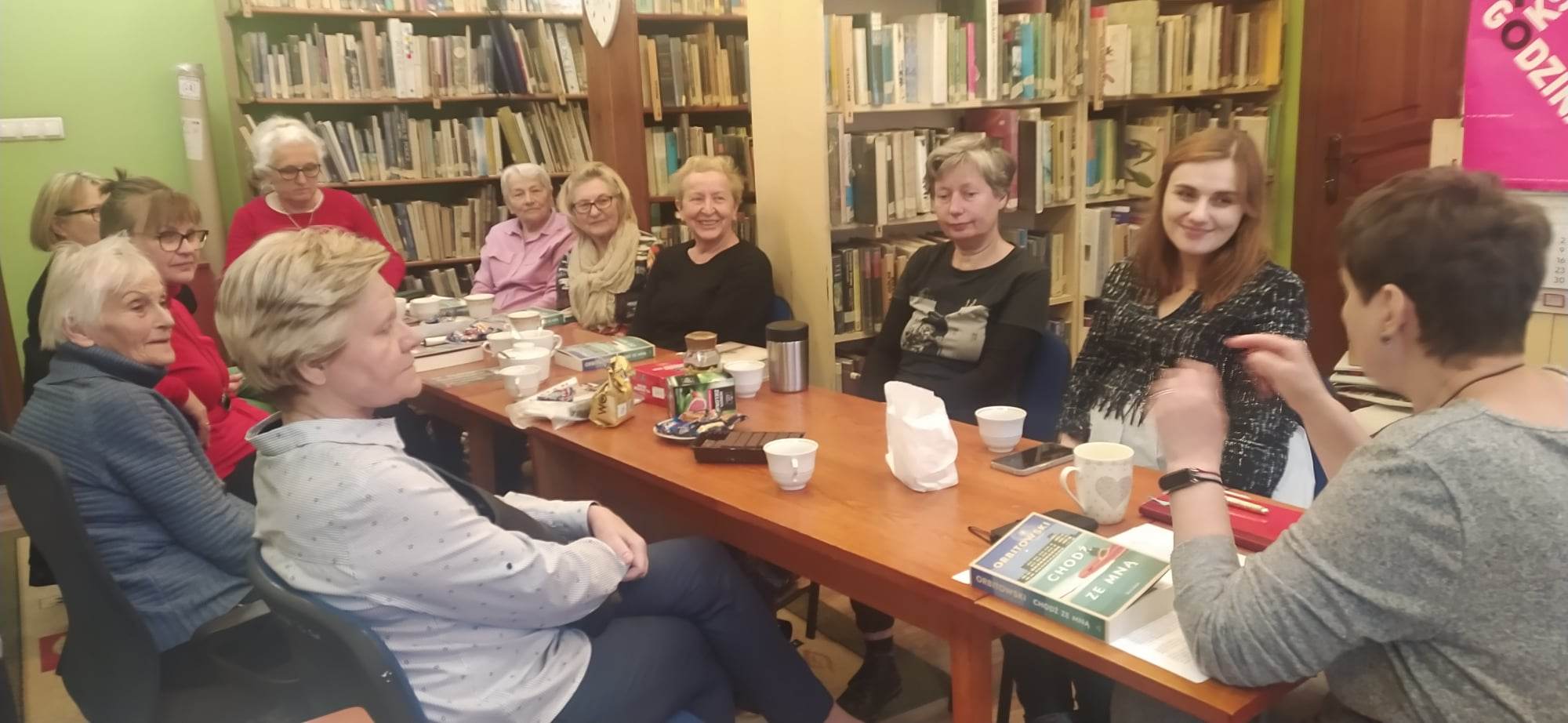 Członkinie Dyskusyjnego Klubu Książki siedzą przy stole i rozmawiają. Część z nich uśmiecha się. Na stole stoją filiżanki z kawą oraz leża ciasteczka.