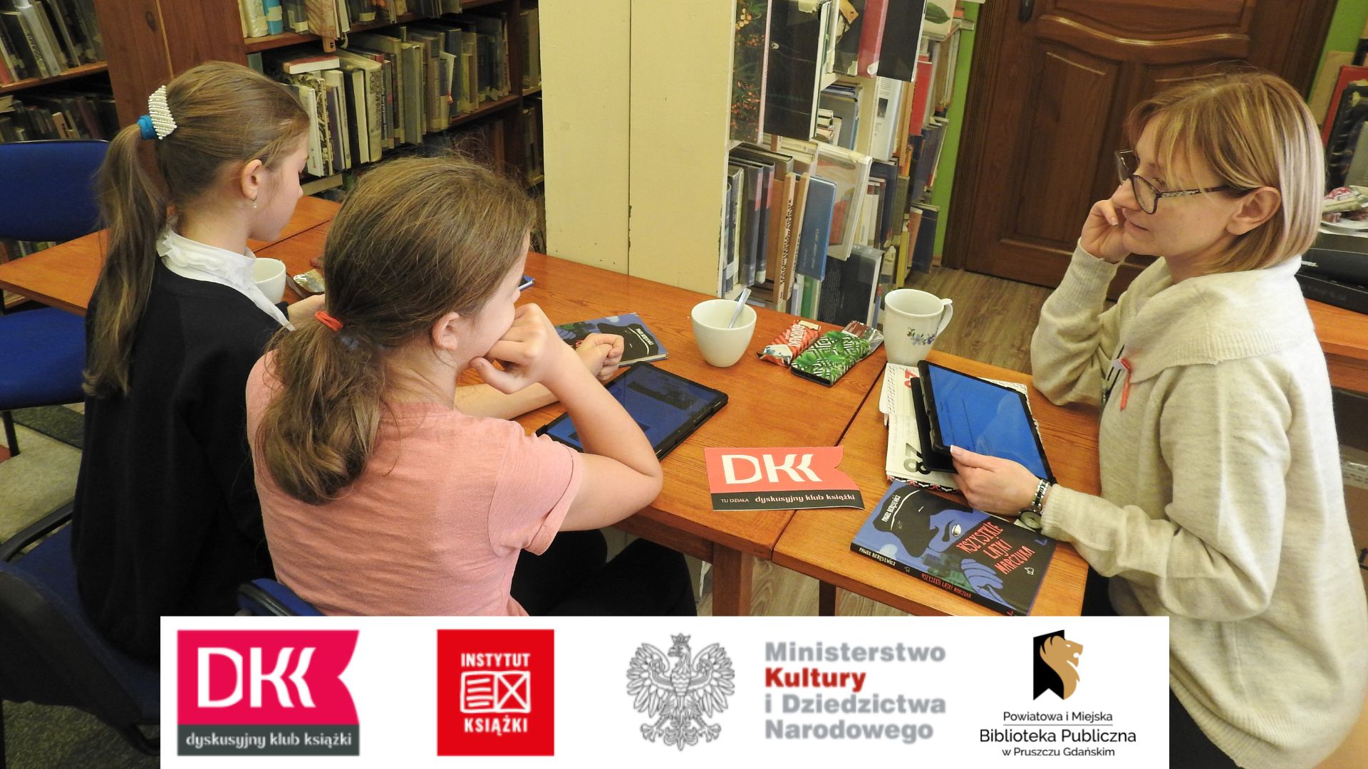 Dwie dziewczynki oraz bibliotekarka, Joanna Łabasiewicz, siedzą przy stole, na którym leży książka pt. Wszystkie lajki Marczuka, logo DKK, kubki oraz tablety. Pani Asia trzyma tablet w dłoniach.