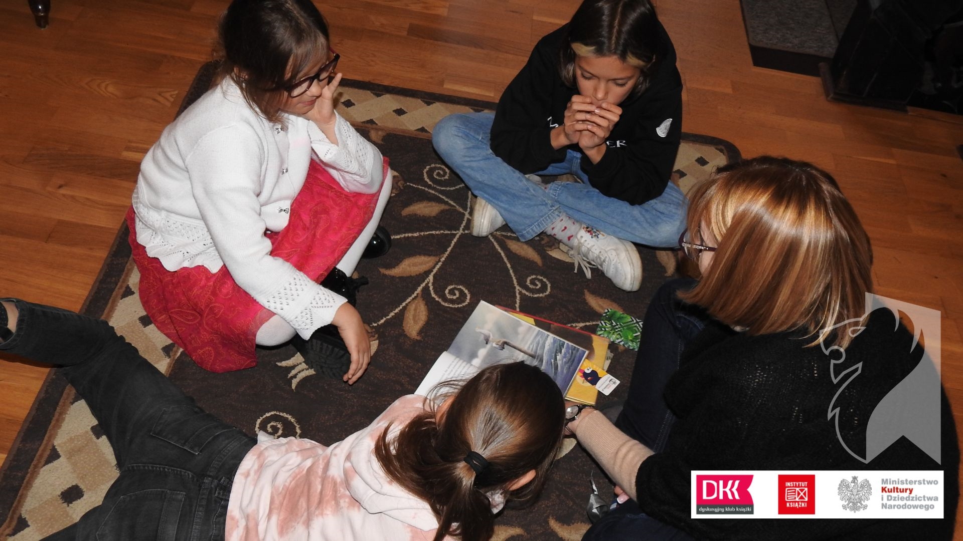 Trójka dzieci oraz moderatorka Dyskusyjnego Klubu Ksiązki siedzą na dywanie. Pośrodku leży otwarta ksiązka.