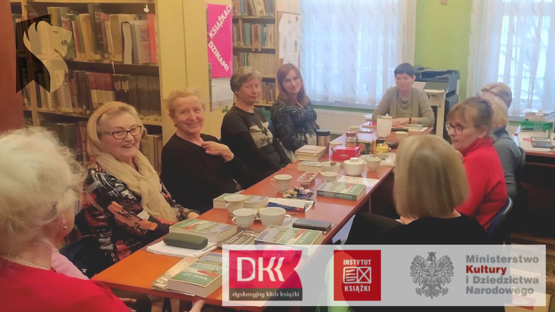 Dziesięć kobiet z Dyskusyjnego Klubu Książk siedzi przy stole, na którym leżą książki oraz stoją filiżanki.