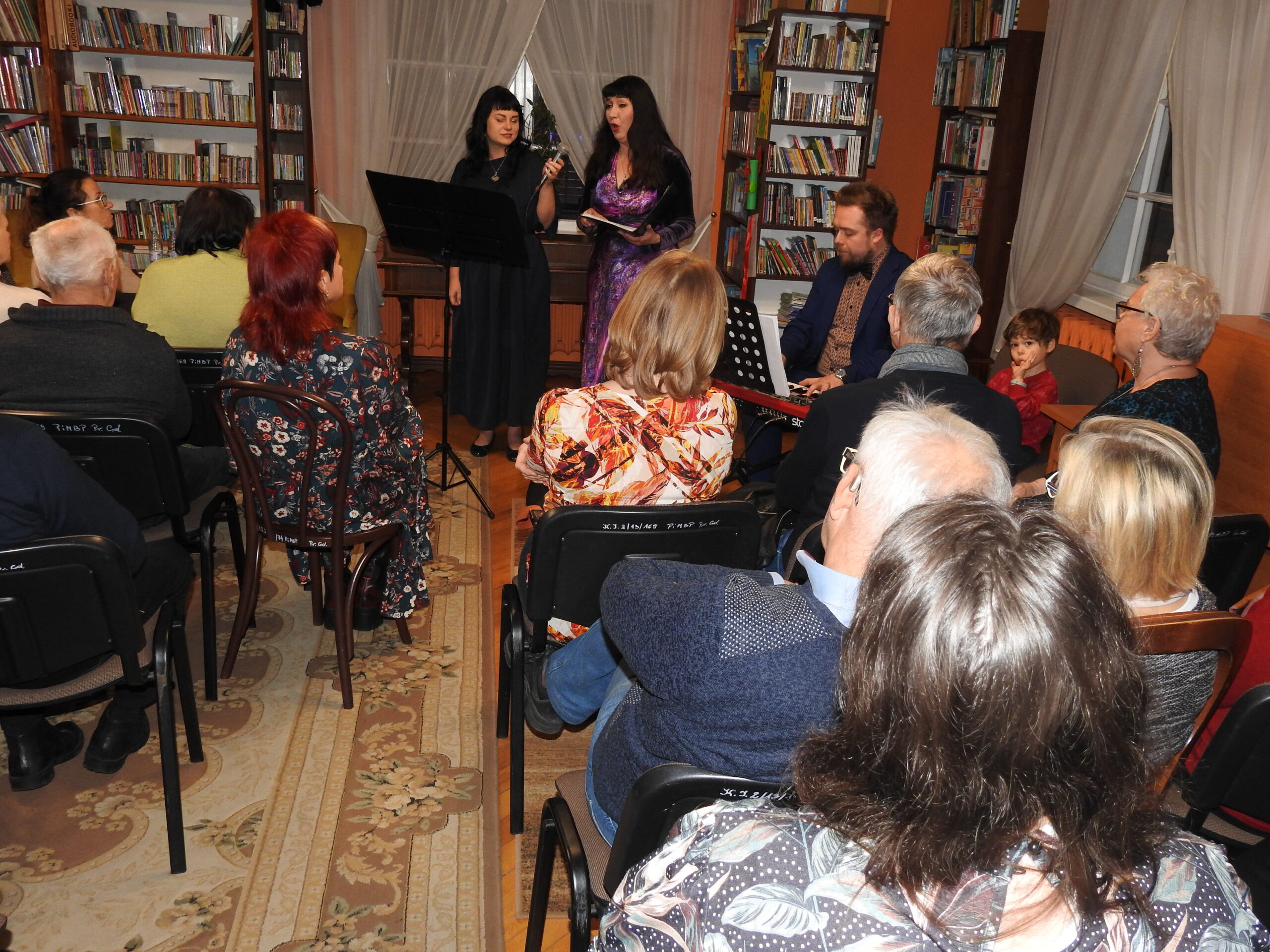 Ewa Jankowska-Mikulska, Kingia Mikulska śpiewają, Andrzej Mikulski gra na przenośnym fortepianie. Przed nimi siedzi publiczność.