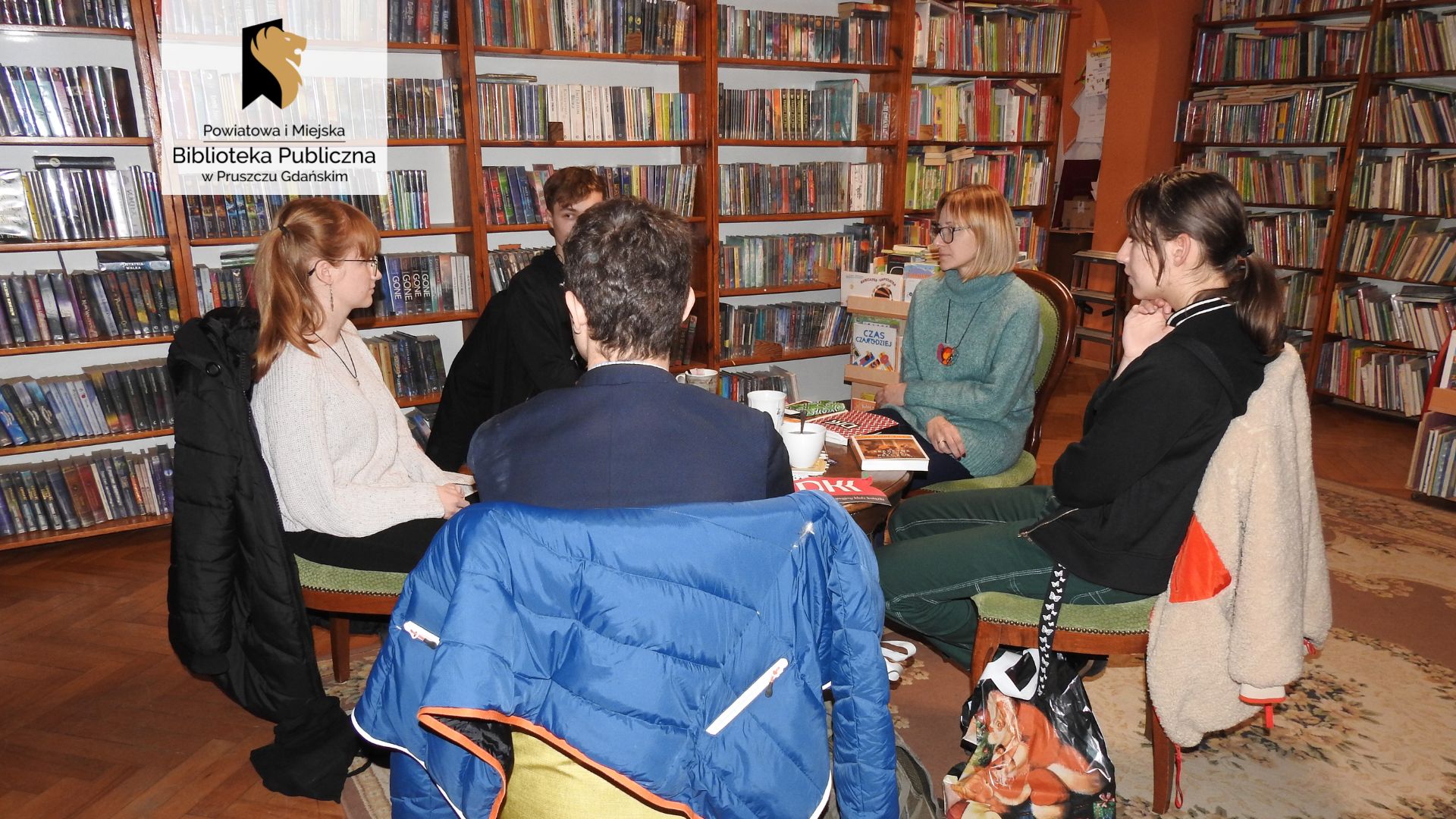 Wokół okrągłego stołu, na krzesłach i fotelach, siedzi 5 osób. Wśród nich jest bibliotekarka Joanna Łabasiewicz. Na stole ksiązki, filiżanki, logo Dyskusyjnego Klubu Książki.