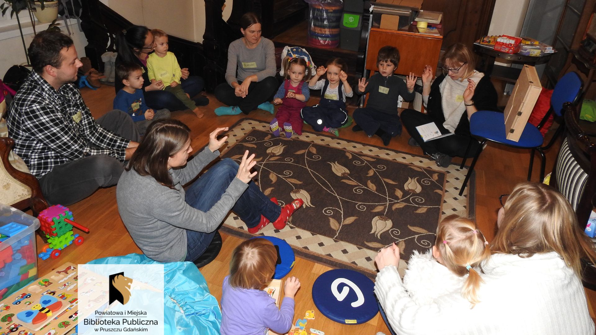 Grupa dzieci oraz kilkoro dorosłych siedzi w dużym kole z bibliotekarką Joanną Łabasiewicz, która na nogach trzyma otwartą książkę. Większość osób ma przed sobą uniesione dłonie pokazując różne gesty.
