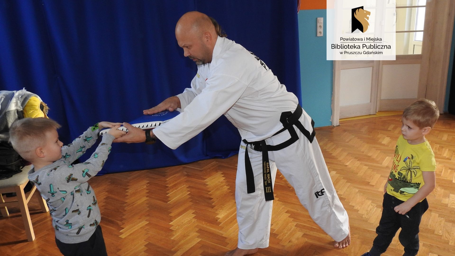 Trener Rafał Mach trzyma worek do ćwiczeń taekwondo wraz z chłopcem, pokazując odpowiednie ułożenie worka. Obok sytuacji przygląda się kolejny chłopiec.