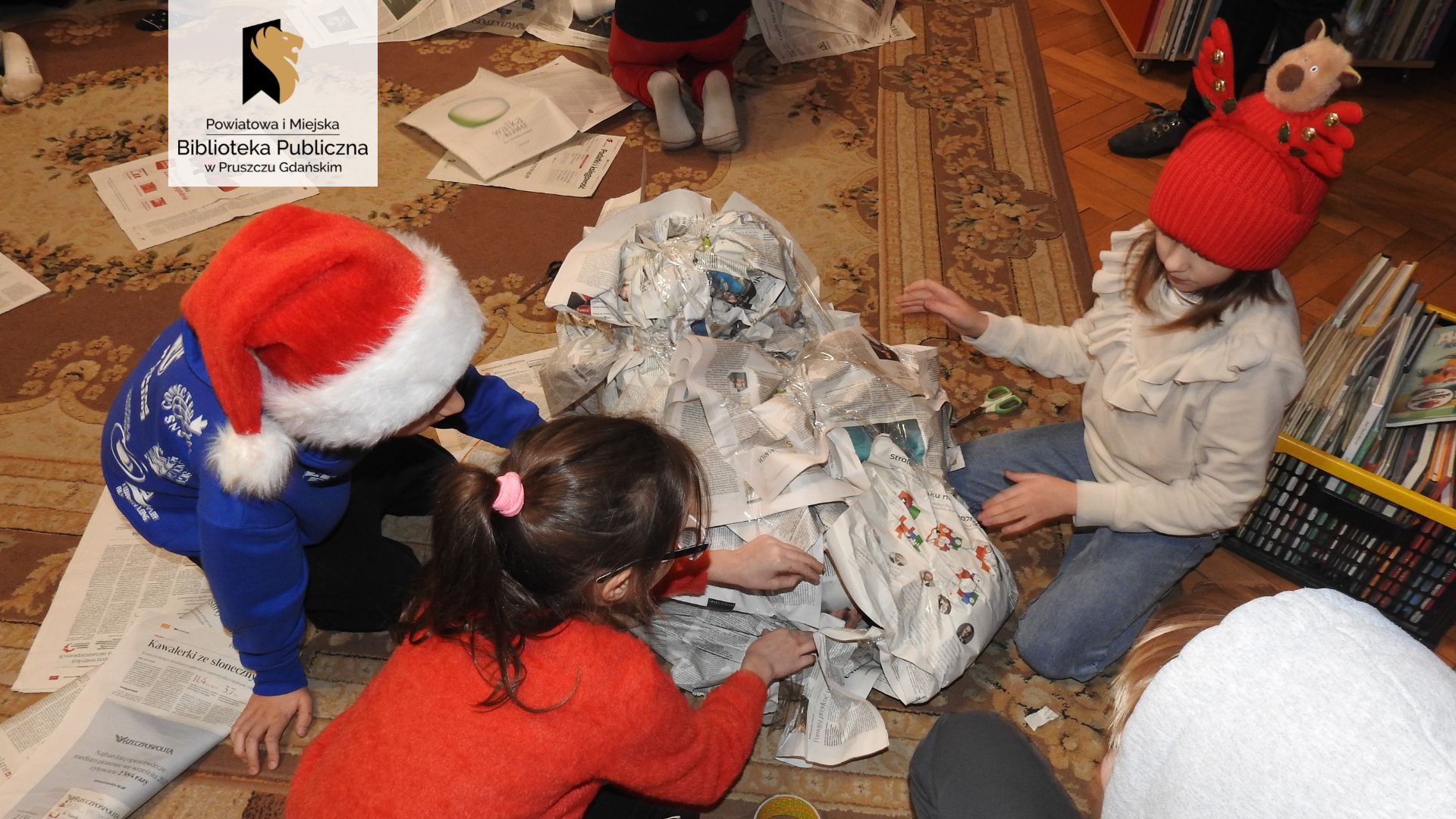 Troje dzieci siedzi wokół dziecka w całości owiniętego w gazety i dokleja kolejne papiery. Wokół rozrzucone strony gazet. Dwoje dzieci ma na głowie świąteczne czapki.