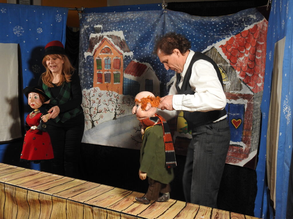 Dwoje aktorów trzyma lalki – mężczyznę i kobietę. W tle zimowa scenografia.