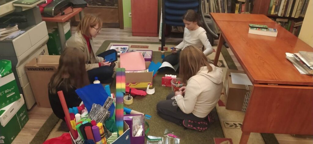 Trzy dziewczynki oraz bibliotekarka, Joanna Łabasiewicz, siedzą na dywanie, na którym rozłożone są kolorowe papiery, taśmy, flamastry. Kobieta trzyma w dłoniach tablet, a dziewczynki materiały plastyczne. Obok dziewczyn stoi duże pudło z kolorowymi bibułami.