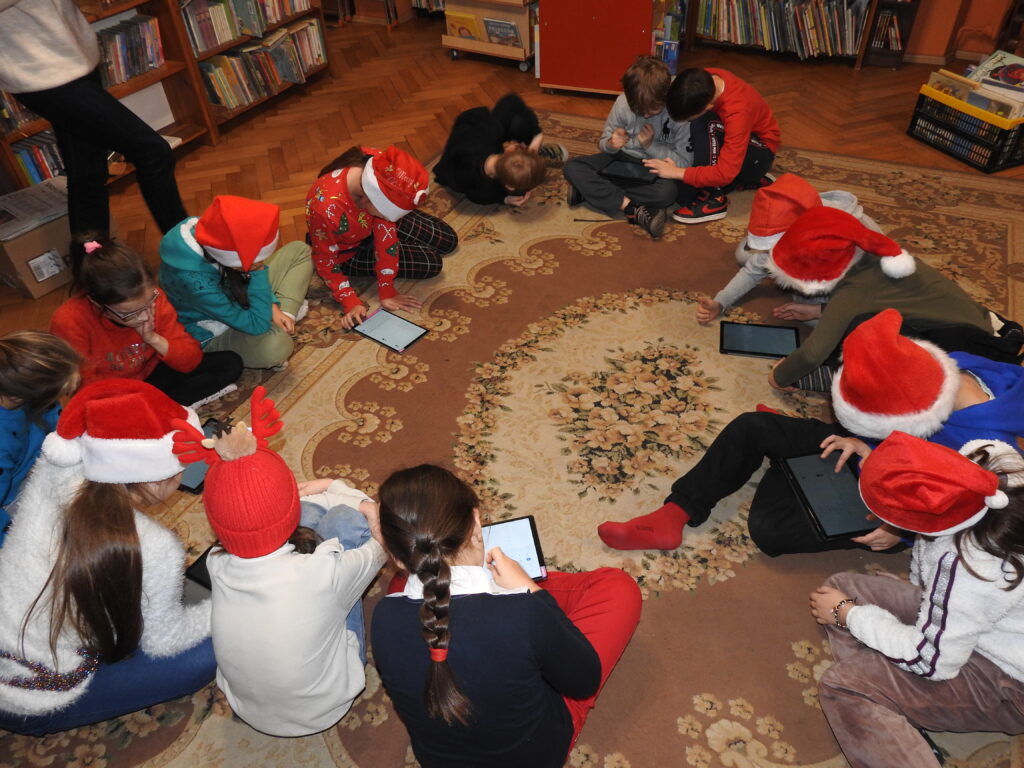 Grupa dzieci siedzi w kole na dywanie i szuka czegoś w tabletach. Wiele dzieci ma na głowach świąteczne czapki.