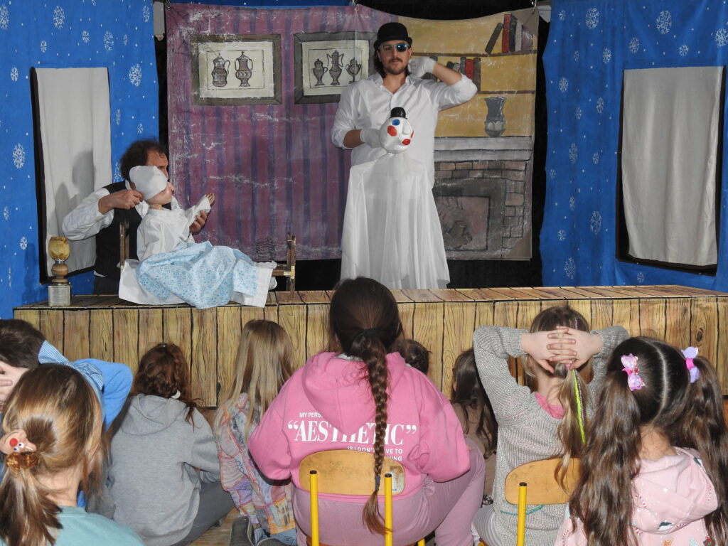 Lalka w piżamie siedzi na łóżku. Podtrzymuje ją aktor. Obok stoi mężczyzna ubrany na biało i trzyma w ręku lalkę ducha. W tle scenografia przedstawiająca wnętrze domu. Naprzeciw grupa dzieci.