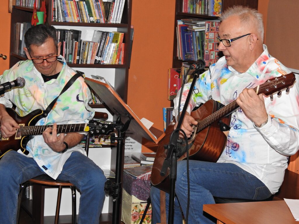Dwóch muzyków siedzi na krzesłach i gra na gitarze. Jeden z nich dodatkowo śpiewa do mikrofonu.