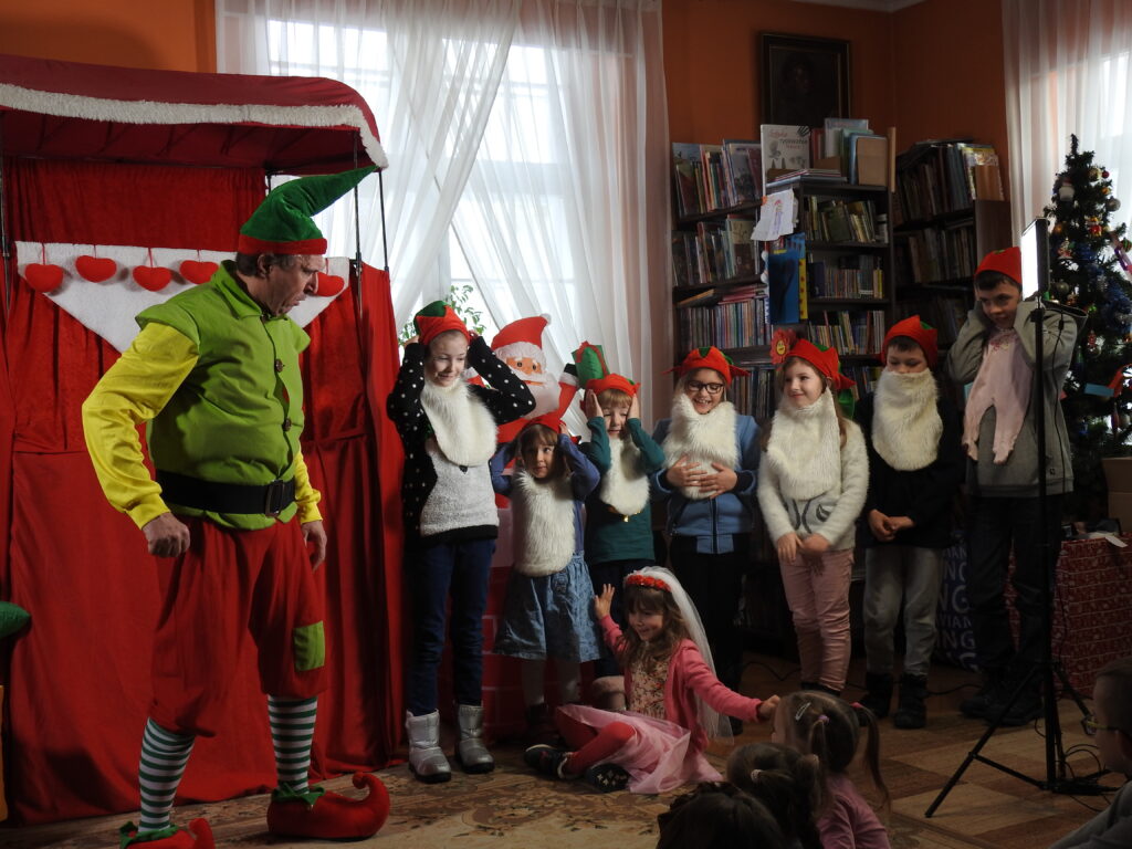 Grupa dzieci ubrana w zielono-czerwone czapki krasnali i brody stoi nad dziewczynką w stroju księżniczki siedzącą na dywanie. Obok nich stoi aktor Teatru Qfer ubrany w strój skrzata. W tle konstrukcja z czerwoną kotarą, regały z książkami, okno.
