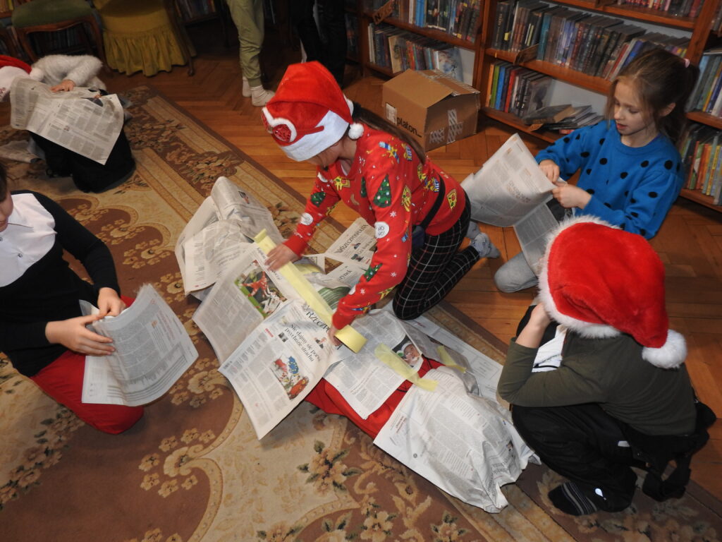 Czworo dzieci klęczy na dywanie wokół dziecka w całości owiniętego w gazety i dokleja kolejne papiery. Dwoje dzieci ma na głowie świąteczne czapki.
