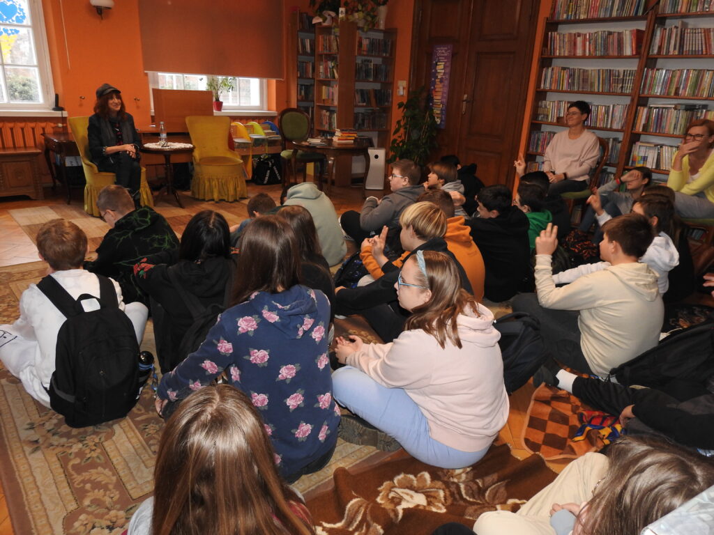 Duża grupa młodzieży siedzi na dywanie i jest zwrócona w stronę Barbary Kosmowskiej siedzącej na fotelu. Kilka osób ma podniesioną rękę do góry. Obok młodzieży, na krzesłach siedzą dwie osoby dorosłe. 