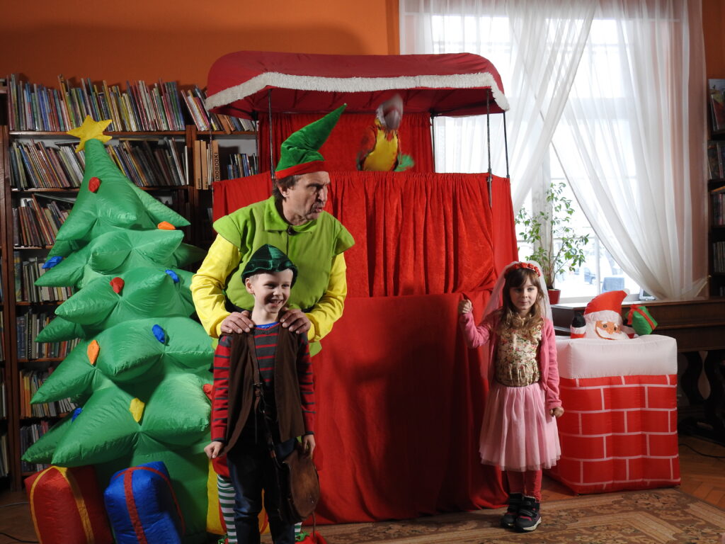 Aktor Teatru Qfer trzyma za ramiona chłopca w stroju myśliwego. Obok nich stoi dziewczynka w stroju księżniczki. Za nimi dmuchana choinka, dmuchany komin z mikołajem oraz konstrukcja pokryta czerwonym materiałem kryjąca zaplecze aktorskie.