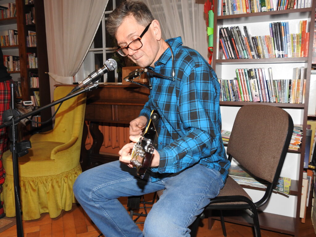 Dmytro Ulyanov siedzi na krześle i gra na gitarze. Na szyi ma zawieszoną harmonię. Przed nim mikrofon.