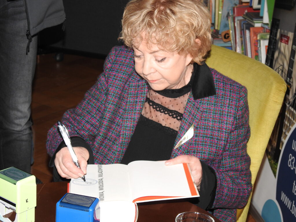 Pani Krystyna Kurczab-Redlich siedzi na fotelu obok stolika i podpisuje książkę pt. Wowa, Wołodia, Władymir.
