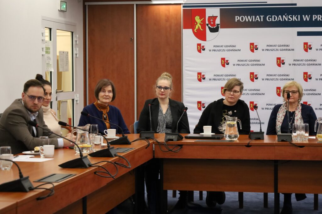 Kilka osób siedzi przy stole, w tym bibliotekarka Milena Jędrzejewska-Kisiel oraz Ela Pozorska z Fundacji KIWI. W tle duży rollup z herbami Powiatu Gdańskiego.