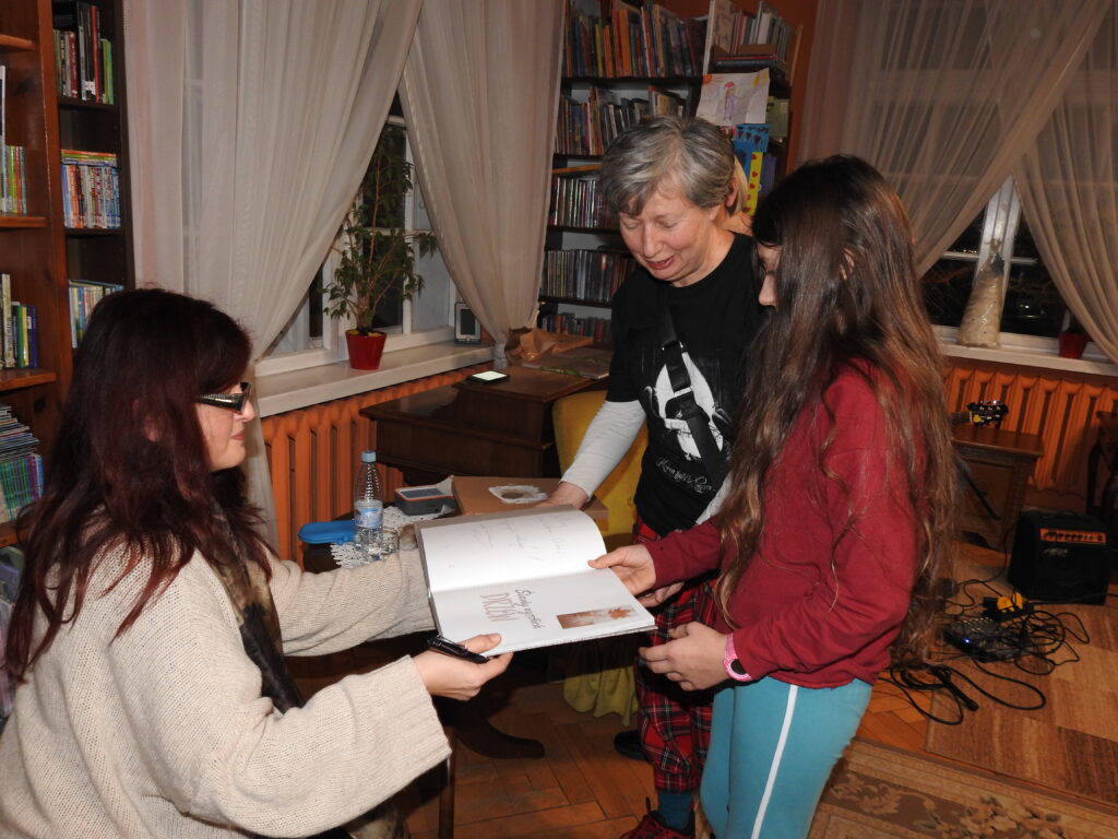 Elena Ulyanova siedzi na fotelu i wręcza otwartą, podpisaną przez siebie książkę dwóm osobom - nastolatce i kobiecie.