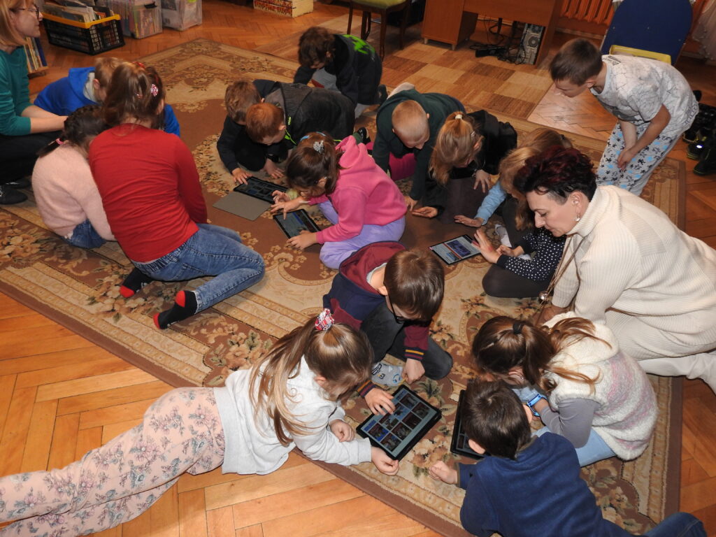 Grupa dzieci siedzi na dywanie i poszukuje informacji na tabletach. 