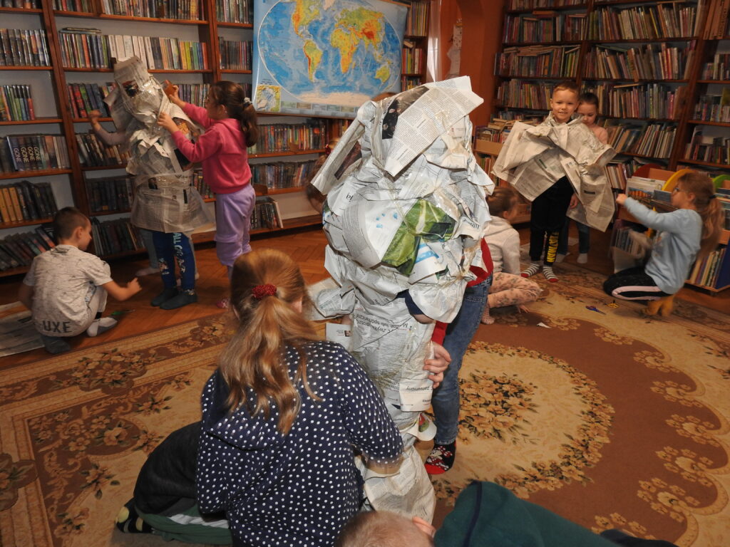 Dzieci są zgromadzone wokół trójki osób owiniętych w gazety. Doklejają kolejne fragmenty papierów. W tle, na regałach z książkami, wisi mapa świata.